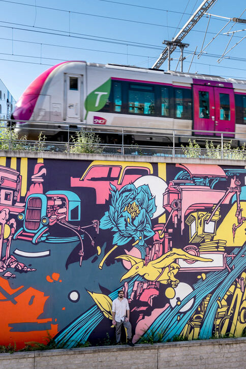 A Street Art Mural paint by Alex Kanos in Paris alex kanos stand on with a graffiti wall painted in background and above there is a railroad with a train